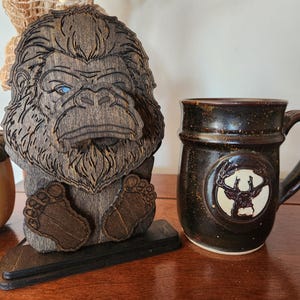 A wooden Bigfoot figurine with detailed laser-cut features, including a stern expression and blue eyes. Beside it, a dark brown ceramic mug with a deer emblem and a speckled finish.