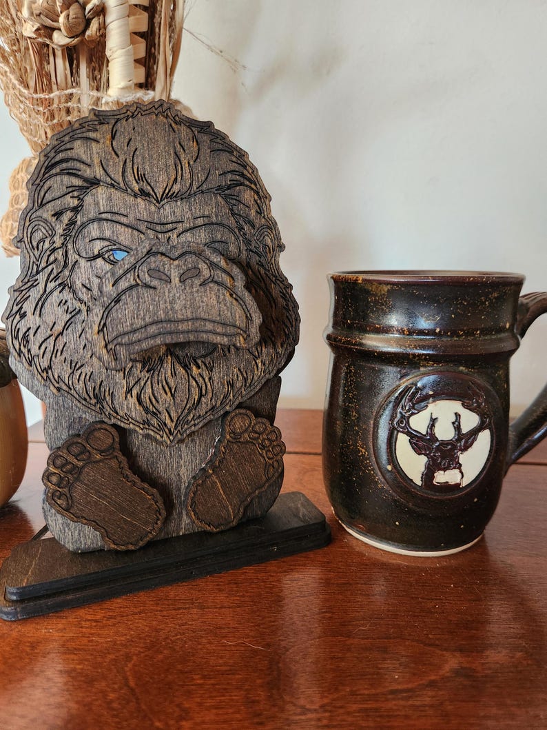 A wooden Bigfoot figurine with detailed laser-cut features, including a stern expression and blue eyes. Beside it, a dark brown ceramic mug with a deer emblem and a speckled finish.