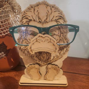 May include: A light-colored wooden eyeglass stand shaped like a gorilla face with detailed laser-etched fur textures. The stand features a protruding nose section designed to hold a pair of dark green plastic-framed eyeglasses securely in place.