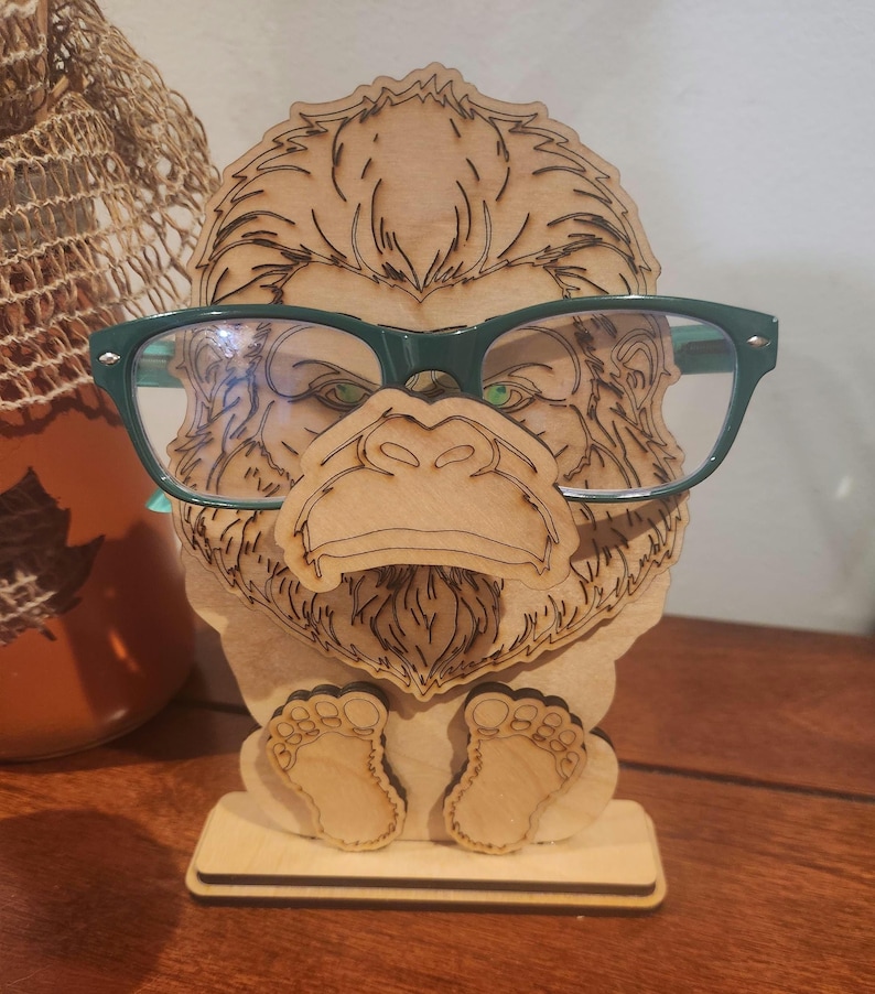 May include: A light-colored wooden eyeglass stand shaped like a gorilla face with detailed laser-etched fur textures. The stand features a protruding nose section designed to hold a pair of dark green plastic-framed eyeglasses securely in place.