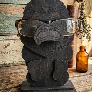 May include: A dark brown wooden eyeglass holder shaped like a gorilla head with a prominent nose and brow. The gorilla features etched fur details and two small feet at the base. A pair of tortoise shell glasses rests on the gorilla's nose.