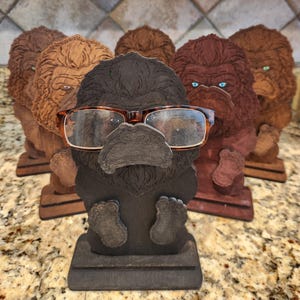 May include: A collection of five wooden gorilla-shaped eyeglass holders in varying shades of brown and black. Each figure features a detailed carved face and a base, with one dark gray gorilla in the foreground holding a pair of tortoiseshell glasses.