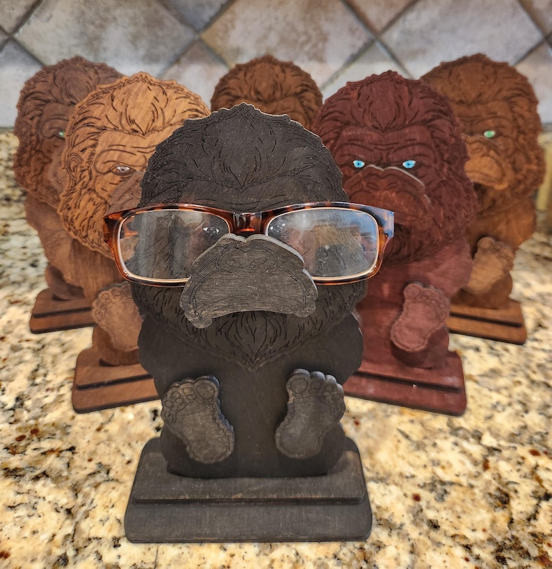 May include: A collection of five wooden gorilla-shaped eyeglass holders in varying shades of brown and black. Each figure features a detailed carved face and a base, with one dark gray gorilla in the foreground holding a pair of tortoiseshell glasses.
