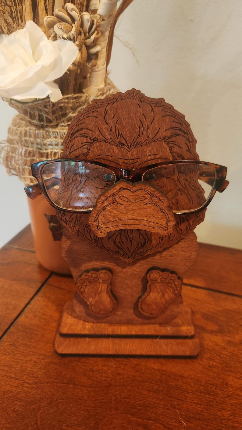 May include: A brown wooden eyeglass holder shaped like a gorilla head with etched facial features. The holder features a flat base and two small feet at the bottom. A pair of brown tortoiseshell-patterned eyeglasses rests on the gorilla's nose.