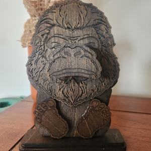 Wooden sculpture of a bigfoot with detailed facial features and feet. The dark brown wood carving is mounted on a rectangular base. The bigfoot's face is intricately carved, showcasing its fur and expression.