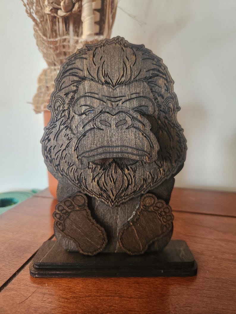 Wooden sculpture of a bigfoot with detailed facial features and feet. The dark brown wood carving is mounted on a rectangular base. The bigfoot's face is intricately carved, showcasing its fur and expression.