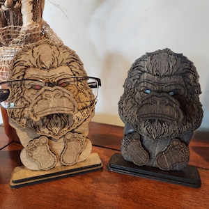 May include: Two wooden gorilla-shaped eyeglass holders standing on rectangular bases. The left holder is light brown with red-tinted eyes, and the right holder is dark brown with blue-tinted eyes. Both feature detailed laser-etched fur textures.