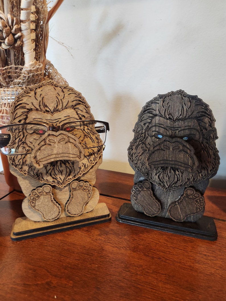 May include: Two wooden gorilla-shaped eyeglass holders standing on rectangular bases. The left holder is light brown with red-tinted eyes, and the right holder is dark brown with blue-tinted eyes. Both feature detailed laser-etched fur textures.