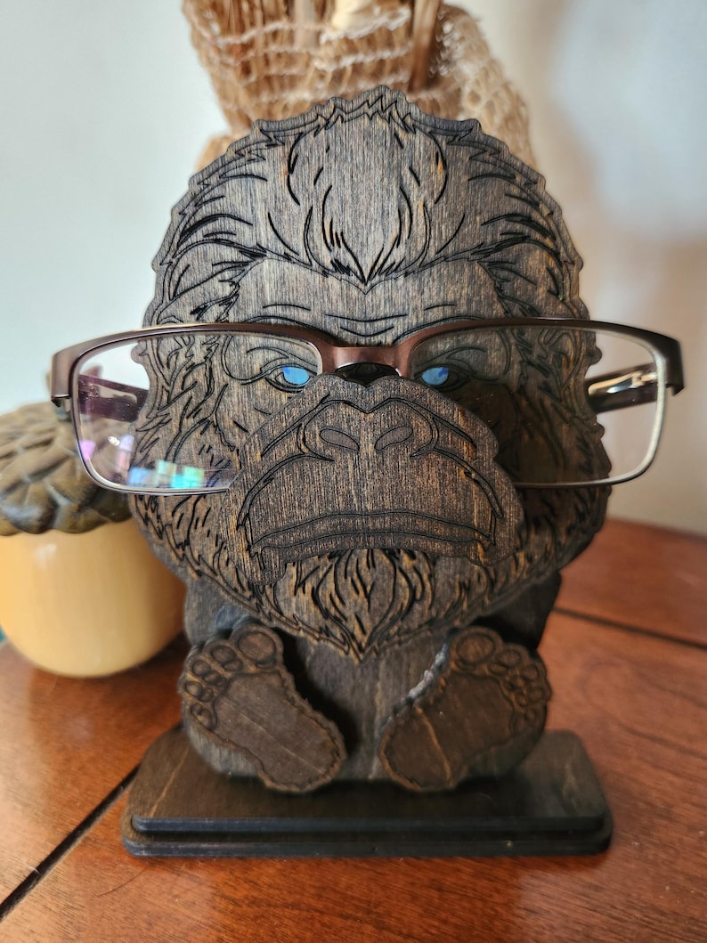 A wooden eyeglass holder shaped like a bigfoot's head, with detailed laser-cut features and feet. The holder has a dark walnut brown finish and is designed to hold a pair of eyeglasses. The bigfoot's eyes are painted blue, and the glasses are brown.