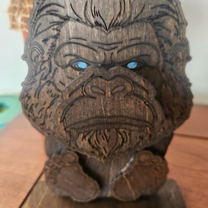 A wooden sculpture of a bigfoot with blue eyes. The bigfoot is carved from dark brown wood, with detailed lines defining its features. The sculpture has a base and is designed to stand upright.
