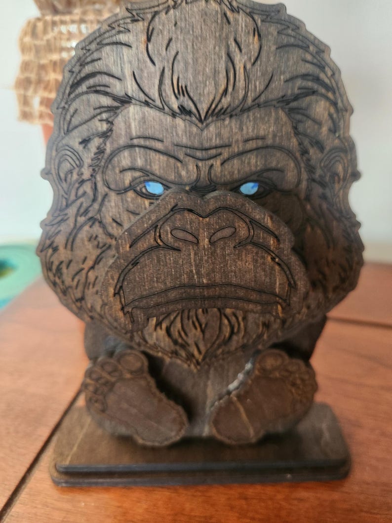 A wooden sculpture of a bigfoot with blue eyes. The bigfoot is carved from dark brown wood, with detailed lines defining its features. The sculpture has a base and is designed to stand upright.
