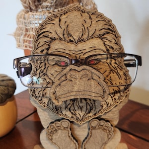 A wooden eyeglass holder shaped like a Bigfoot with red eyes. The holder is made of light brown wood and has a detailed laser-cut design. The Bigfoot is wearing a pair of black-framed glasses. The holder has a small base and two feet.