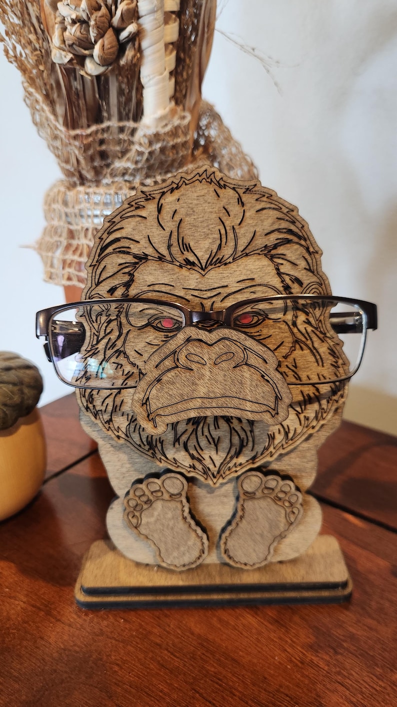 A wooden eyeglass holder shaped like a Bigfoot with red eyes. The holder is made of light brown wood and has a detailed laser-cut design. The Bigfoot is wearing a pair of black-framed glasses. The holder has a small base and two feet.