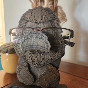 A wooden eyeglass holder shaped like a Bigfoot with glasses. The holder is made of dark brown wood and has detailed carvings of the gorilla's face, feet, and body. The glasses are brown-framed.