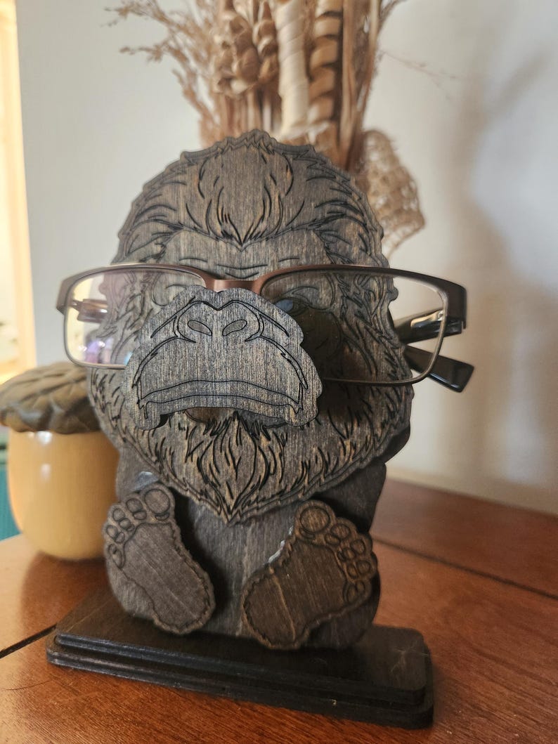 A wooden eyeglass holder shaped like a Bigfoot with glasses. The holder is made of dark brown wood and has detailed carvings of the gorilla's face, feet, and body. The glasses are brown-framed.
