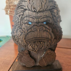 A wooden sculpture of a bigfoot with blue eyes. The bigfoot is carved from dark brown wood and has detailed features. The sculpture is sitting on a wooden base.