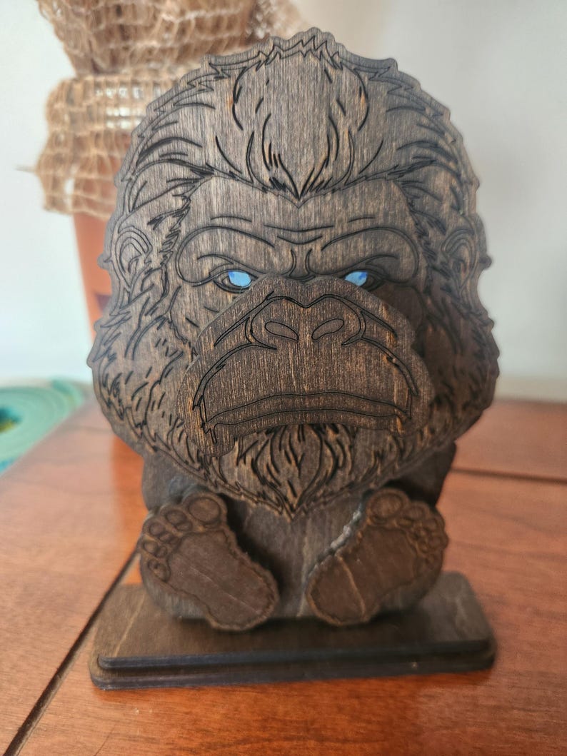 A wooden sculpture of a bigfoot with blue eyes. The bigfoot is carved from dark brown wood and has detailed features. The sculpture is sitting on a wooden base.
