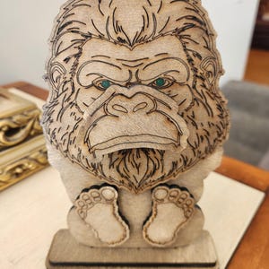 May include: A laser-cut wooden stand shaped like a gorilla with a prominent face, textured fur details, and two small feet at the base. The gorilla has dark etched lines defining its features and two small, circular green accents for its eyes.