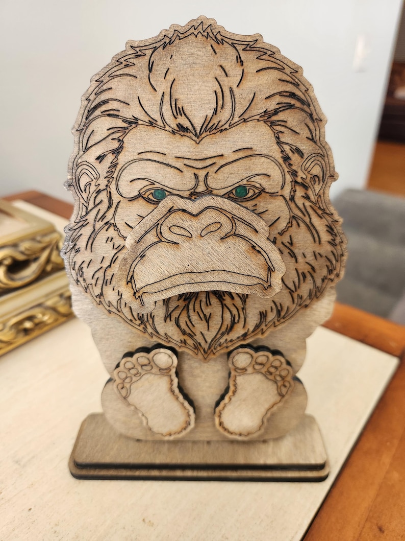 May include: A laser-cut wooden stand shaped like a gorilla with a prominent face, textured fur details, and two small feet at the base. The gorilla has dark etched lines defining its features and two small, circular green accents for its eyes.