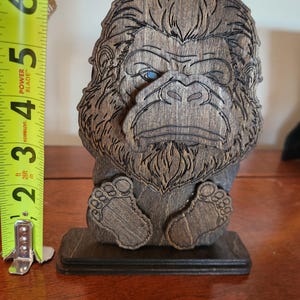 A wooden Bigfoot sculpture with intricate carved details and a dark walnut stain finish. The sculpture features a detailed face with blue eyes and is approximately 6 inches tall.