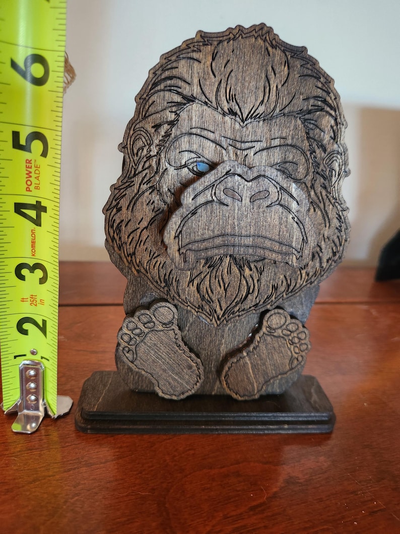 A wooden Bigfoot sculpture with intricate carved details and a dark walnut stain finish. The sculpture features a detailed face with blue eyes and is approximately 6 inches tall.