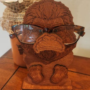 May include: A brown wooden eyeglass holder shaped like a gorilla head with etched facial features. The holder features a flat base and two small feet at the bottom. A pair of brown tortoiseshell-patterned eyeglasses rests on the gorilla's nose.