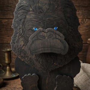 May include: A dark brown wooden gorilla sculpture featuring engraved fur details and two bright blue circular eyes. The gorilla is depicted in a seated position with its feet visible at the bottom, resting on a flat rectangular wooden base.