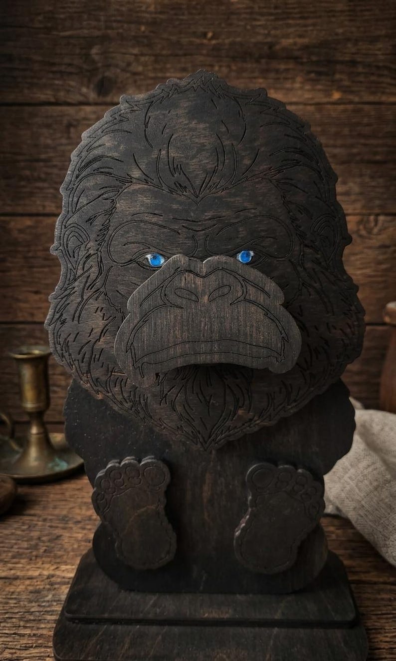 May include: A dark brown wooden gorilla sculpture featuring engraved fur details and two bright blue circular eyes. The gorilla is depicted in a seated position with its feet visible at the bottom, resting on a flat rectangular wooden base.