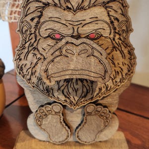 May include: A laser-cut wooden figurine of a gorilla with a detailed face, prominent brow, and textured fur. The gorilla has bright red eyes and is depicted in a seated position with its feet visible at the bottom, mounted on a rectangular base.