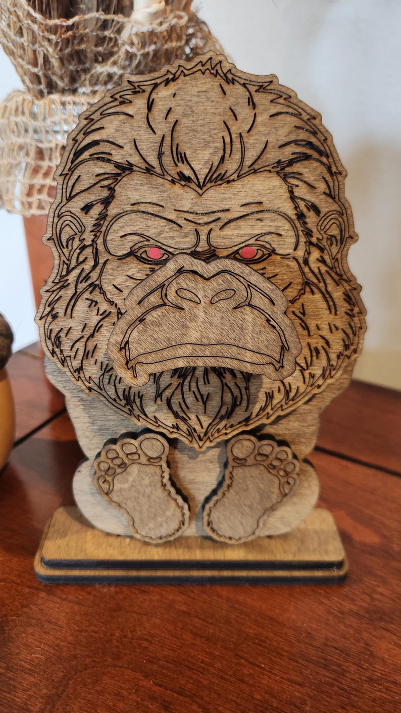 May include: A laser-cut wooden figurine of a gorilla with a detailed face, prominent brow, and textured fur. The gorilla has bright red eyes and is depicted in a seated position with its feet visible at the bottom, mounted on a rectangular base.
