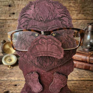 May include: A dark purple wooden eyeglass holder shaped like a gorilla head with a prominent nose and feet at the base. The gorilla features engraved fur details and is wearing a pair of brown tortoiseshell-patterned eyeglasses across its face.