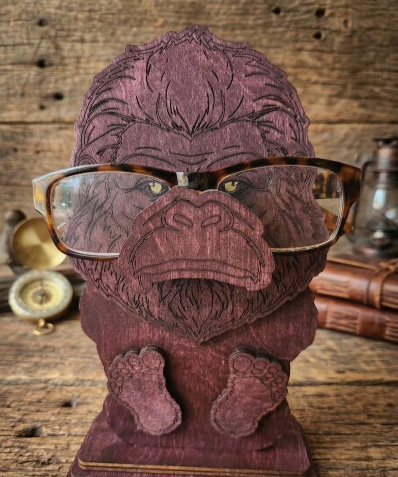 May include: A dark purple wooden eyeglass holder shaped like a gorilla head with a prominent nose and feet at the base. The gorilla features engraved fur details and is wearing a pair of brown tortoiseshell-patterned eyeglasses across its face.