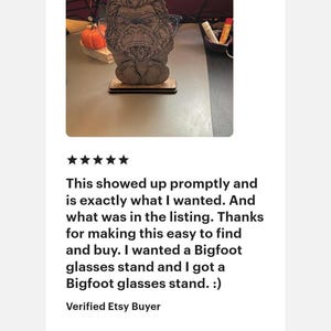 May include: A brown, laser-cut wooden glasses stand shaped like a Bigfoot figure with detailed fur texture. The figure is standing on a rectangular base and is currently holding a pair of thin-framed eyeglasses across its face area.
