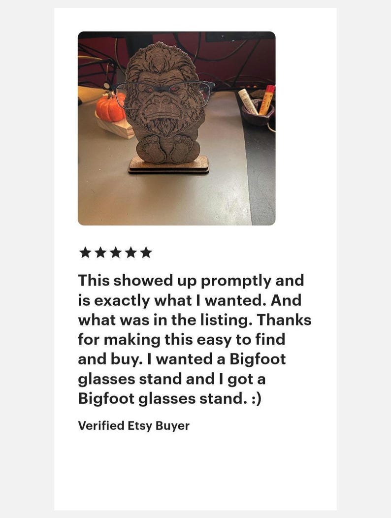 May include: A brown, laser-cut wooden glasses stand shaped like a Bigfoot figure with detailed fur texture. The figure is standing on a rectangular base and is currently holding a pair of thin-framed eyeglasses across its face area.