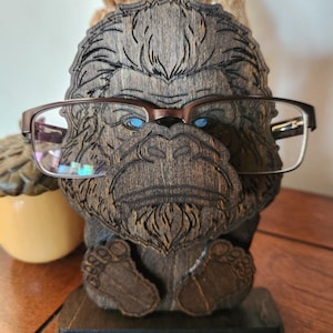 A wooden eyeglass holder shaped like a Bigfoot with glasses resting on its face. The holder is brown with intricate carved details, including the Bigfoot's face, hair, and feet. The glasses have a brown frame and clear lenses. The holder sits on a rectangular base.