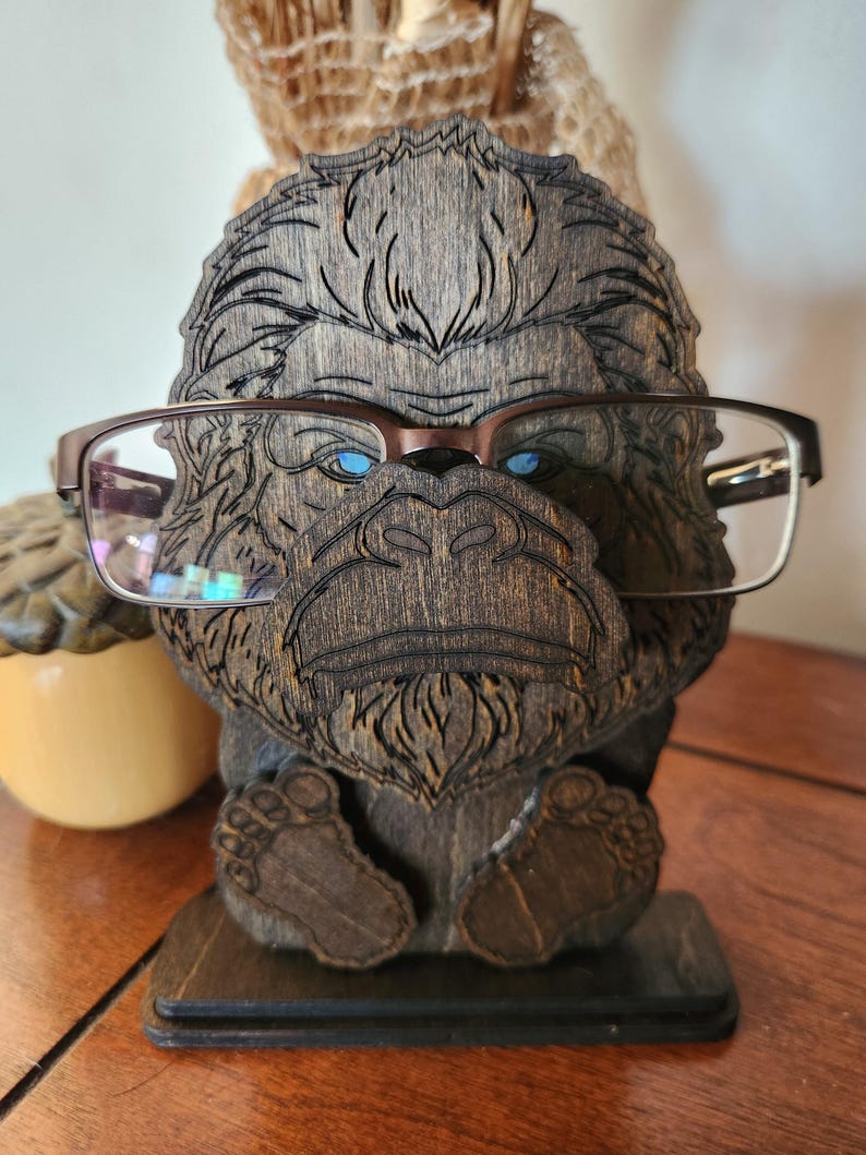 A wooden eyeglass holder shaped like a Bigfoot with glasses resting on its face. The holder is brown with intricate carved details, including the Bigfoot's face, hair, and feet. The glasses have a brown frame and clear lenses. The holder sits on a rectangular base.