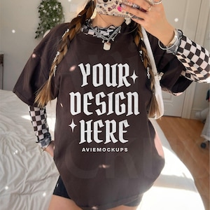 Könnte beinhalten: Ein braunes Oversize-T-Shirt mit dem Text "YOUR DESIGN HERE" und "AVIEMOCKUPS" auf der Vorderseite. Das Shirt wird von einer Person mit langen geflochtenen Haaren getragen.