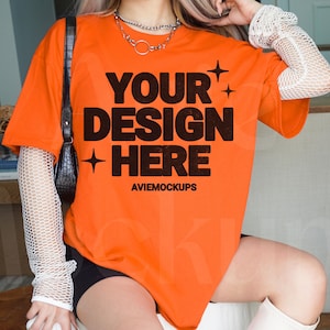 Puede incluir: Camiseta naranja de gran tamaño con el texto "YOUR DESIGN HERE" y las palabras "AVIEMOCKUPS" debajo. La camiseta la lleva puesta una persona con el pelo largo y rubio.
