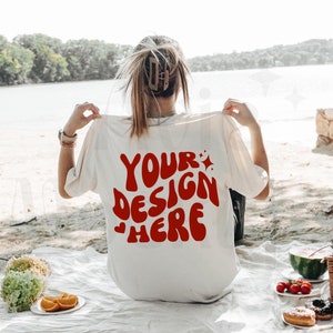 以下が含まれることがあります： レトロなフォントで赤色の「YOUR DESIGN HERE」というテキストが入ったクリーム色のTシャツ。白いブランケットの上に座っている人がシャツを持ち上げており、前景にはピクニックが広がっています。