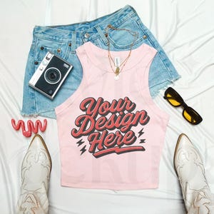 May include: A light pink tank top with the text "Your Design Here" in red and black script, paired with denim shorts, white cowboy boots, a vintage camera, sunglasses, and jewelry. The outfit is arranged on a white surface.