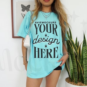 May include: A light blue oversized t-shirt with the text "AVIEMOCKUPS YOUR design HERE" printed in black. The shirt is being worn by a person with long blonde hair.