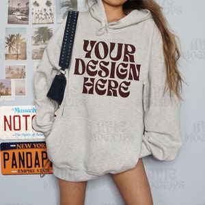 Puede incluir: Una sudadera con capucha gris claro con el texto "YOUR DESIGN HERE" en una fuente de color rojo oscuro. La sudadera con capucha tiene un bolsillo delantero y una capucha con cord&oacute;n. Un bolso negro con una correa con tachuelas cuelga del hombro. Las matr&iacute;culas y las fotos de la playa est&aacute;n en la pared.