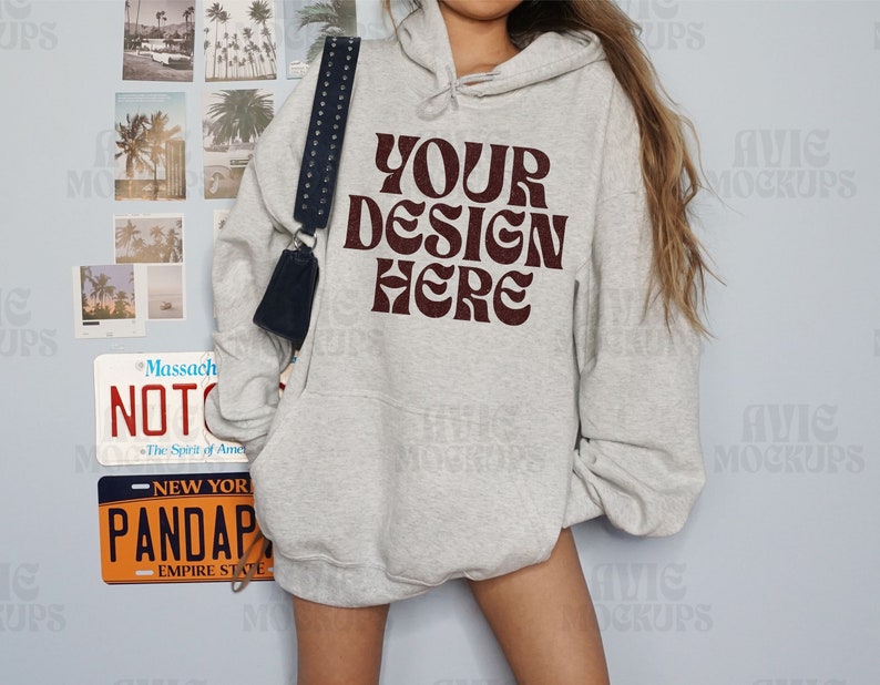 Puede incluir: Una sudadera con capucha gris claro con el texto "YOUR DESIGN HERE" en una fuente de color rojo oscuro. La sudadera con capucha tiene un bolsillo delantero y una capucha con cord&oacute;n. Un bolso negro con una correa con tachuelas cuelga del hombro. Las matr&iacute;culas y las fotos de la playa est&aacute;n en la pared.