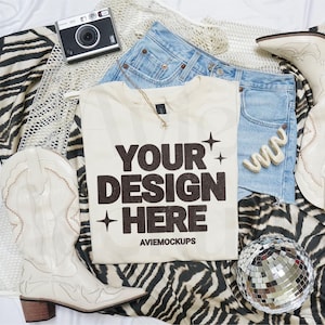 Puede incluir: Una camiseta blanca con el texto "YOUR DESIGN HERE" en letras negras. La camiseta está colocada sobre una tela de estampado de cebra con una bola de discoteca, botas de vaquero y unos pantalones cortos de mezclilla.
