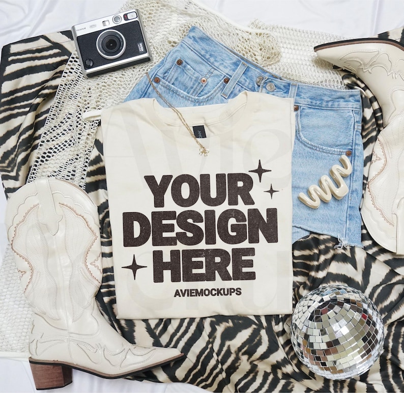 May include: A white t-shirt with the text "YOUR DESIGN HERE" in black lettering. The shirt is laid out on a zebra print fabric with a disco ball, cowboy boots, and a pair of denim shorts.