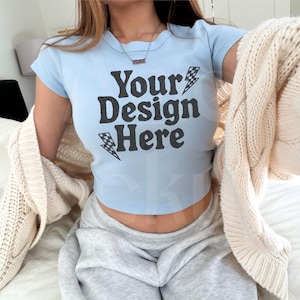 May include: Light blue crop top with the text "Your Design Here" in black. The shirt has a crew neck and short sleeves. A cream-colored cardigan is draped over the shoulders. The person is wearing gray sweatpants.
