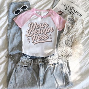 May include: A flat lay featuring a white and pink short-sleeved t-shirt with the text "Your Design Here", light-wash denim jeans, white sunglasses, a newspaper, and a net bag. A silver chain belt is on the jeans. The items are arranged on a white sheet.
