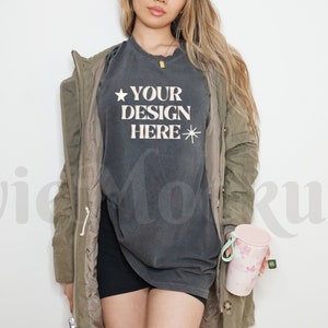 May include: A woman wearing a dark grey t-shirt with the text "YOUR DESIGN HERE" and a green jacket. She is holding a pink and white tumbler.