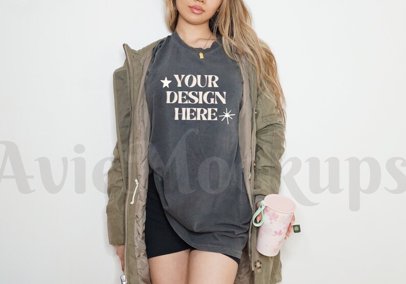 May include: A woman wearing a dark grey t-shirt with the text "YOUR DESIGN HERE" and a green jacket. She is holding a pink and white tumbler.