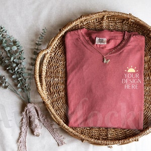 K&ouml;nnte beinhalten: Ein rotes T-Shirt mit einem wei&szlig;en Sonnenmotiv und dem Text "YOUR DESIGN HERE" auf einem Weidenkorb.
